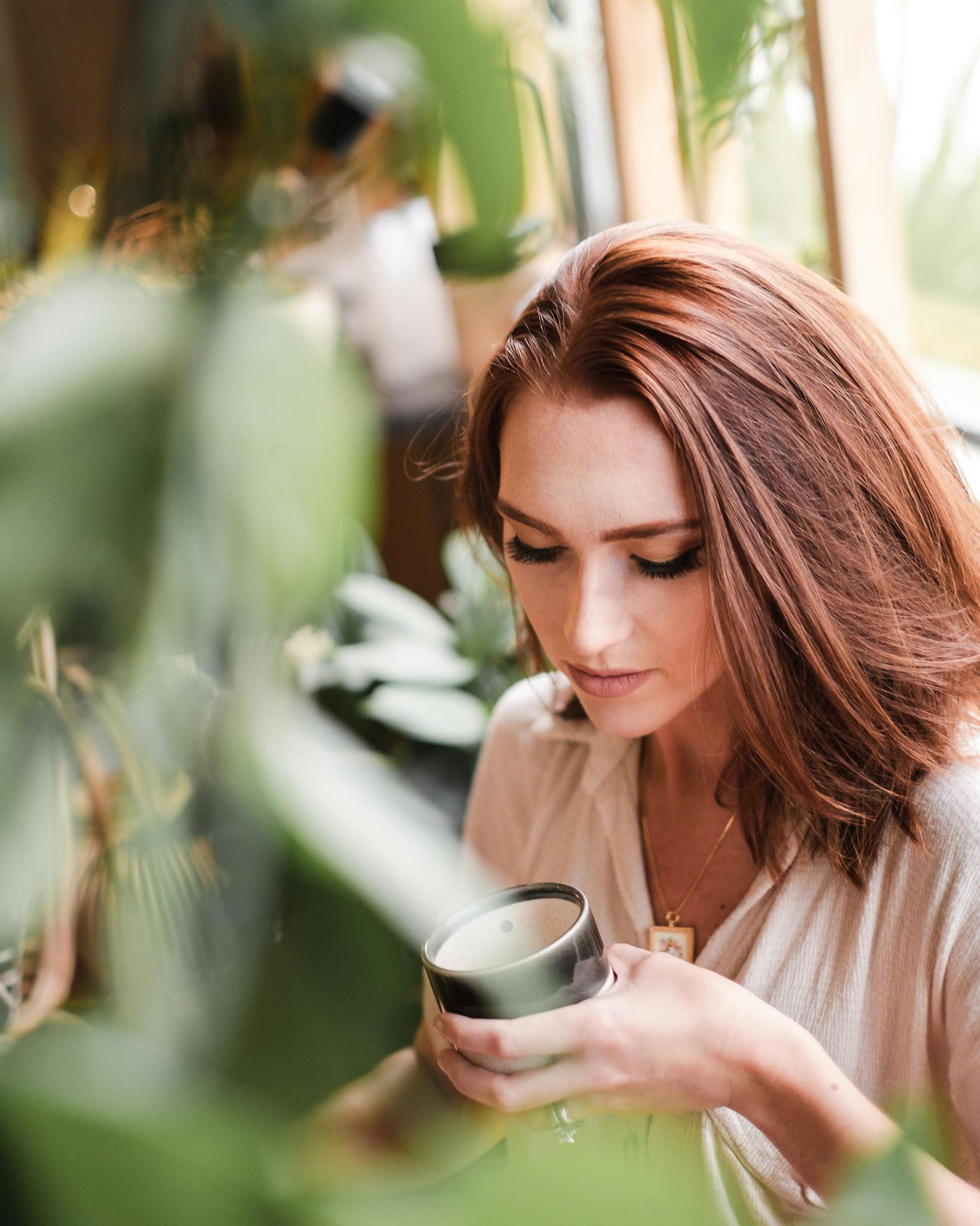 A serene moment of a woman enjoying a hot beverage surrounded by lush greenery indoors.