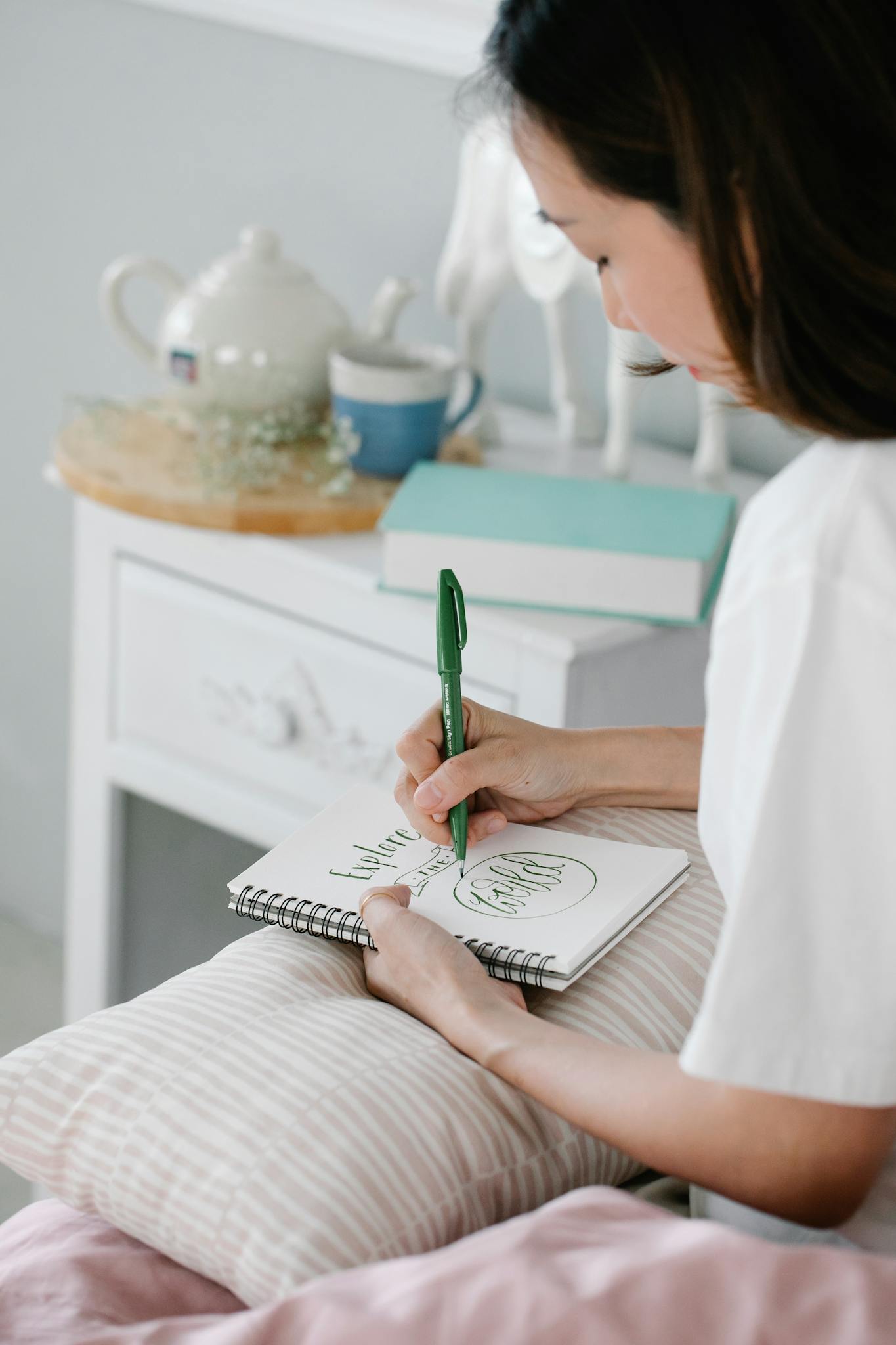A woman sits comfortably on a bed, writing in a notebook, surrounded by a cozy bedroom setup.
